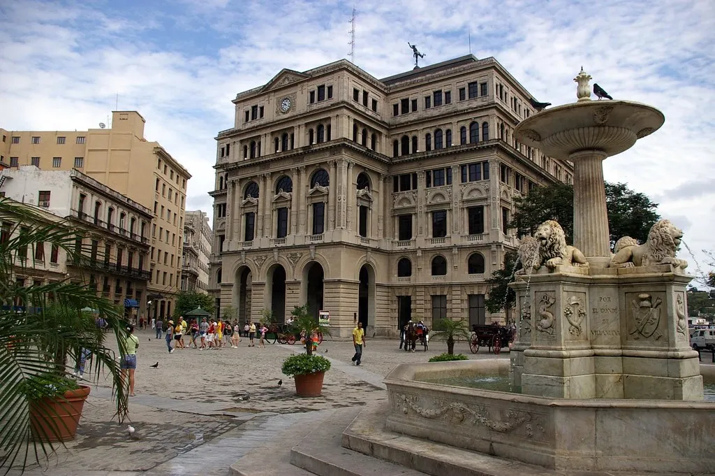 Una fuente con leones que preside la plaza, un edificio de estilo neoclásico de fondo y gente paseando