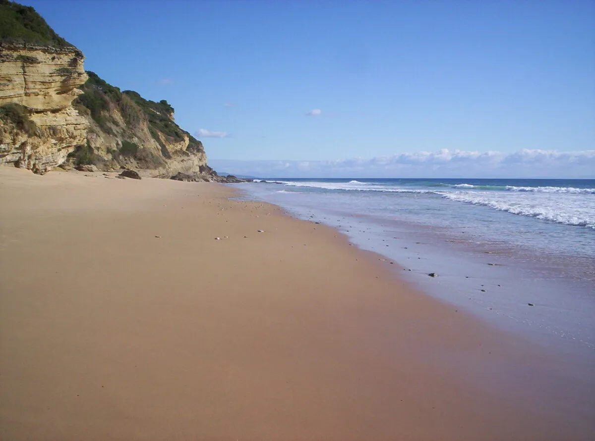 Panorámica de la playa durante un día soleado con arena dorada, aguas cristalinas y zona rocosa al fondo