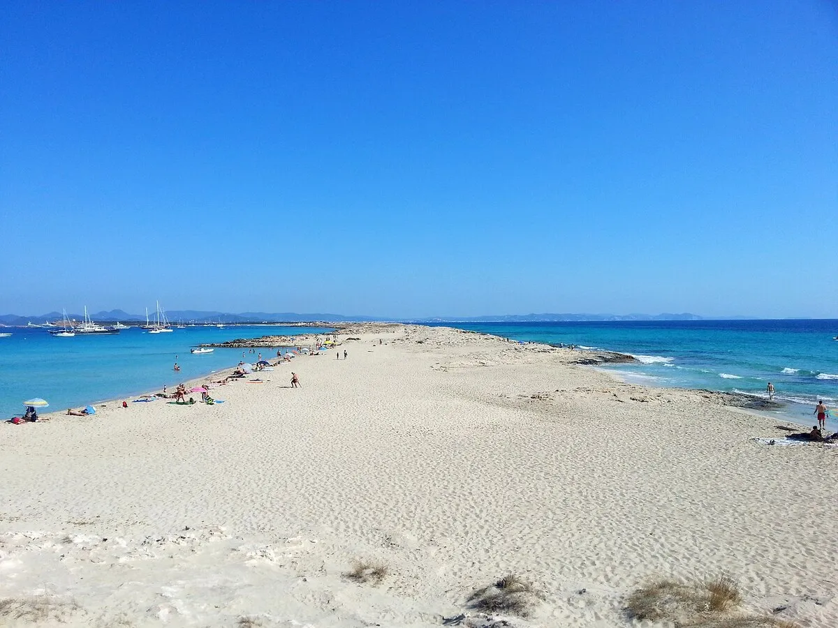 El istmo sobre el que se encuentra la playa con gente con toallas tomando el sol y barcos de vela sobre las aguas turquesas