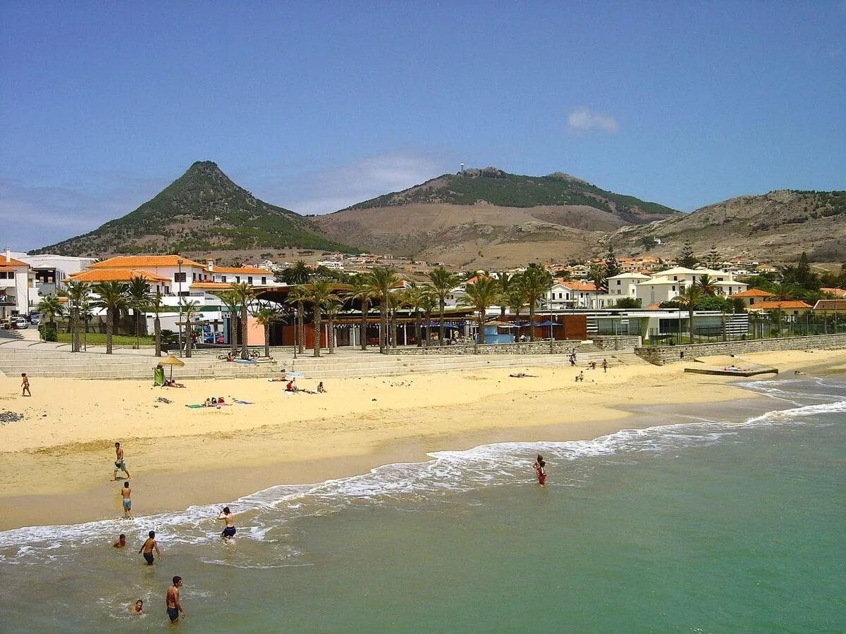 Costa de una playa de la isla con palmeras en el paseo marítimo, montañas al fondo y gente bañándose en el mar