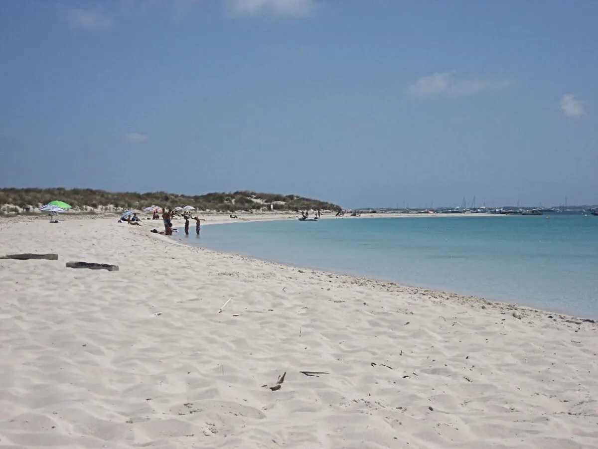 Playa de arena blanca y aguas cristalinas con gente tomando el sol y barcos de vela de fondo