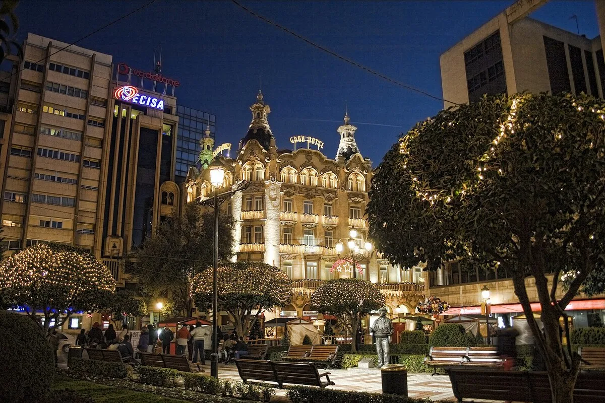 La plaza con luces de navidad sobre los arboles y un edificio neoclásico de fondo
