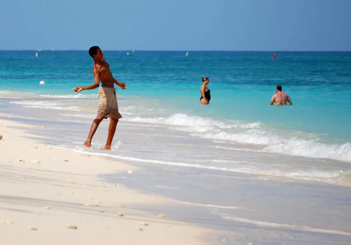 Personas bañándose en el mar color turquesa y un chico en la orilla de la playa de arena blanca