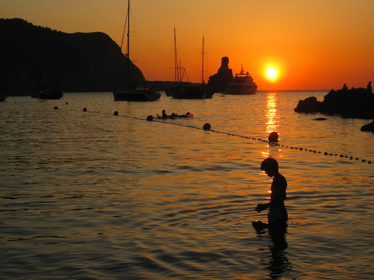 Algunos barcos atracados en la cala, un chico bañándose junto a las bollas y un precioso atardecer de fondo.