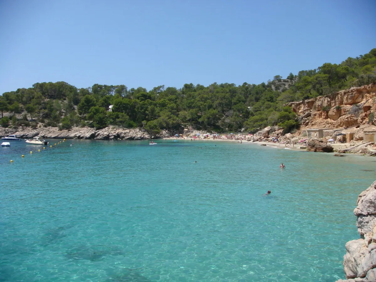 El agua cristalina y la arena dorada de la cala con gente y sombrillas de fondo durante un día soleado