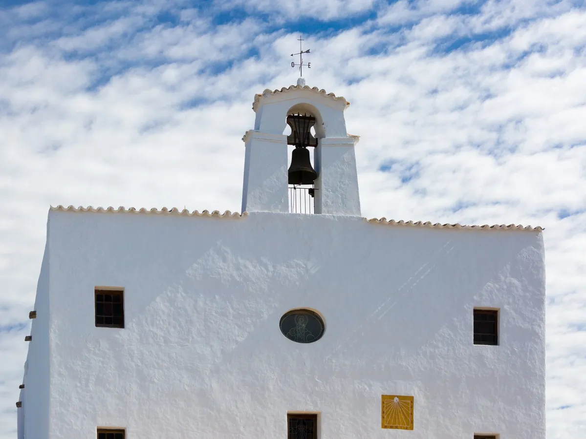 La fachada principal de Iglesia de color blanco y con detalles amarillos. En la parte de arriba hay una campana que preside.