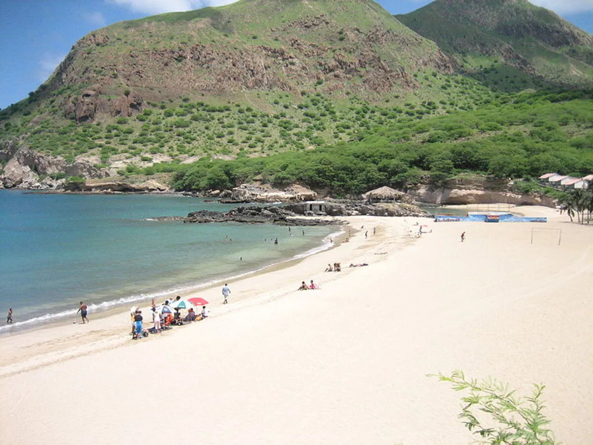 Panorámica de la playa con arena blanca y con la montaña repleta de vegetación. También hay gente con sombrillas en la orilla